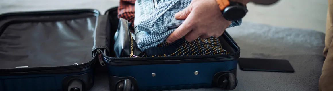 Un homme prépare sa valise de cabine avec des vêtements, un carnet de notes et des écouteurs.